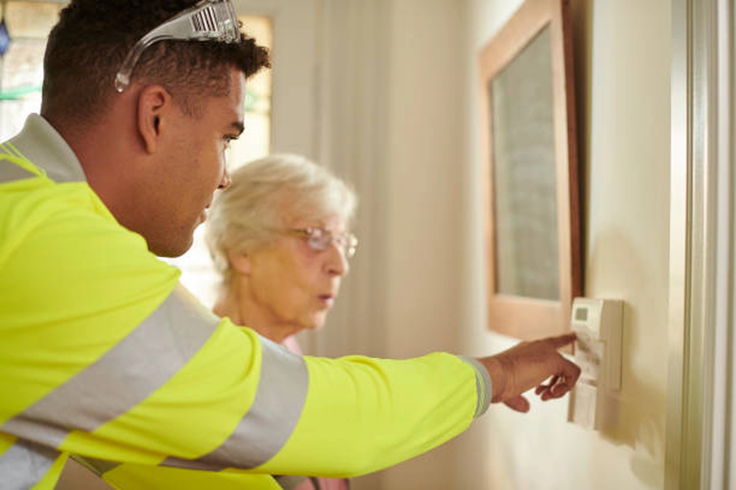 Pötschke Elektro - Elektrofachunternehmen - Ein Klimatechniker in einer Warnweste zeigt einer älteren Dame, wie man ein an der Wand montiertes Thermostat bedient.
